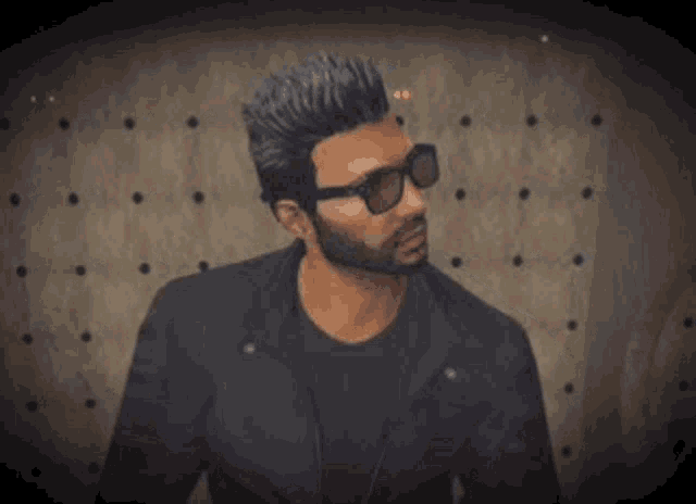 a man with a beard and sunglasses is wearing a black jacket and black shirt .
