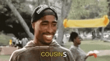 a man wearing a hat and a hoodie is smiling and saying `` cousins '' while standing in a park .