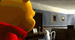 winnie the pooh is standing in a living room next to a man sitting on a couch .