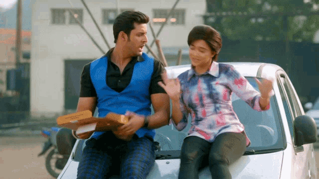 a man and a woman are sitting on the hood of a car and the man is holding a pizza box