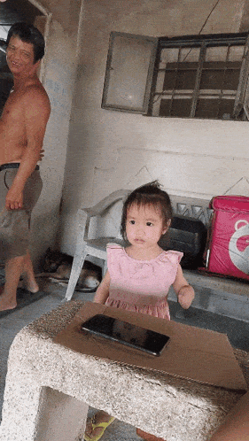 a little girl in a pink dress is sitting at a table with a cell phone on it