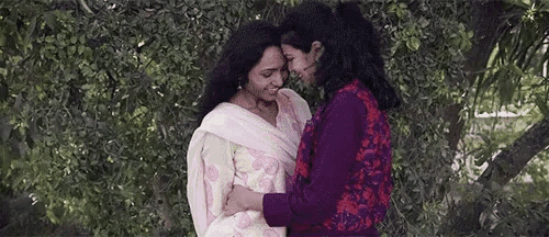 two women are hugging each other in front of a tree in a park .