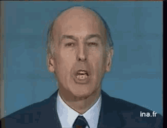 a bald man in a suit and tie is giving a speech on a television screen .
