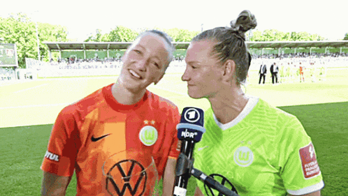 two women are standing next to each other on a soccer field and one has a microphone with the number 1 on it .