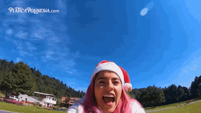 a woman with pink hair is wearing a santa hat with the website platicapolynesia.com in the background