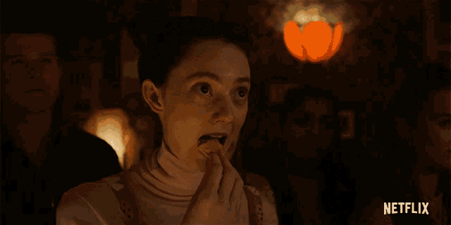 a woman in a pink turtleneck stands in a dark room with a netflix logo on the bottom right