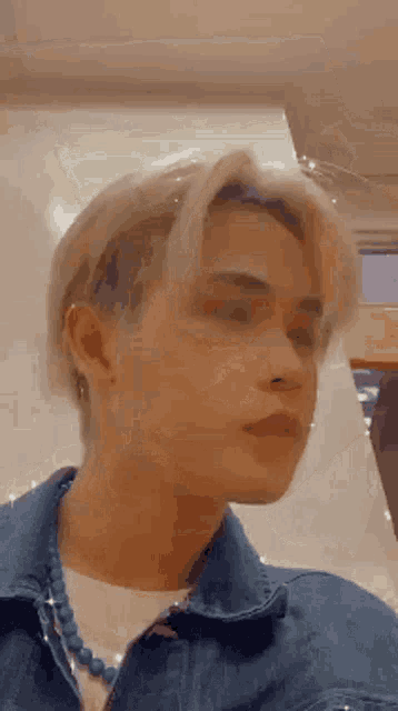 a young man wearing a denim jacket and a blue necklace is looking at the camera .