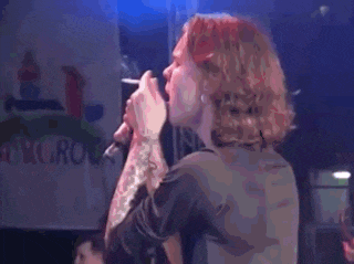 a man with long blonde hair is singing into a microphone in front of a sign that says ' burger '