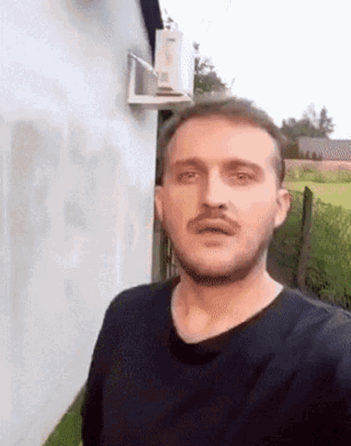 a man with a mustache is standing in front of a white wall with a fan on it .