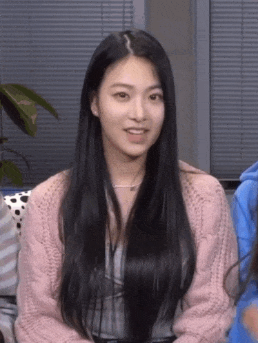 a woman with long black hair is wearing a pink sweater and smiling while sitting on a couch .