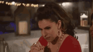 a woman in a red dress and earrings is sitting at a table with her hands folded in prayer .