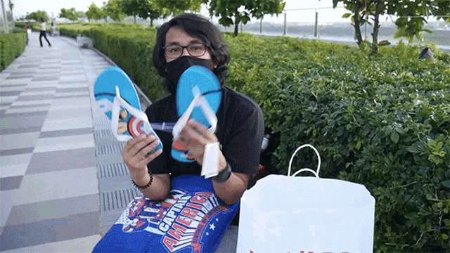 a man wearing a mask holds up a pair of flip flops next to a bag that says captain america