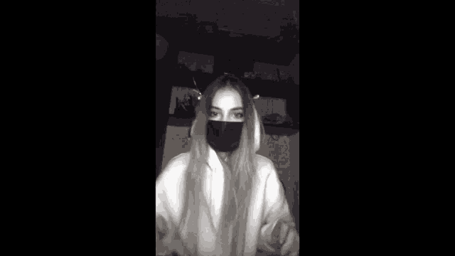a black and white photo of a woman wearing a black mask and giving a peace sign .