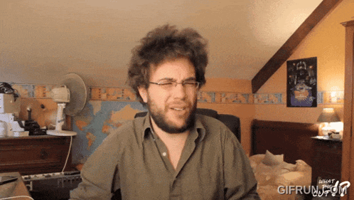 a man with glasses and a beard is sitting in front of a keyboard and a poster that says what the gif