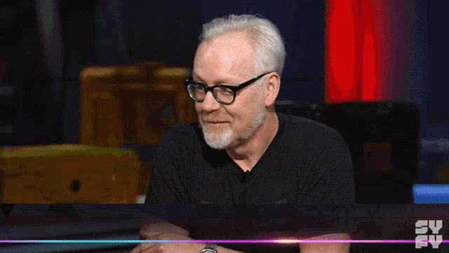 a man with glasses and a beard is sitting in front of a tv screen that says sy