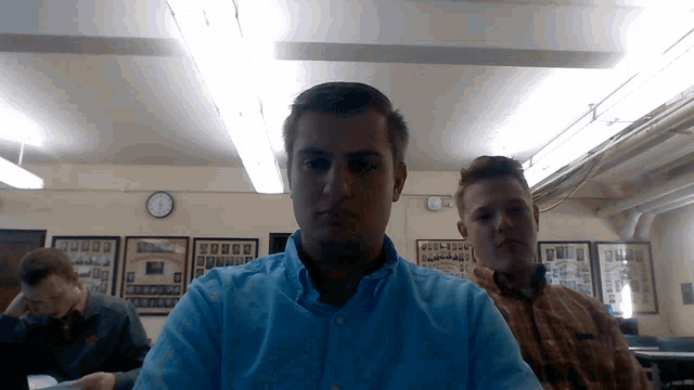 a man in a blue shirt looks at the camera in a room with a clock on the wall
