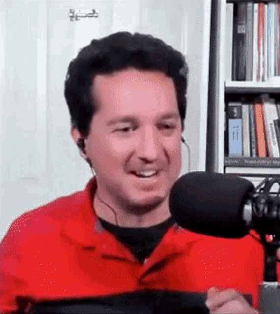 a man in a red shirt is talking into a microphone in front of a bookshelf