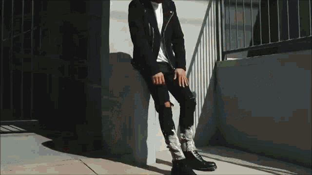 a man is sitting on a wall wearing a black leather jacket and ripped jeans .