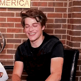 a young man is smiling in front of a brick wall with a sign that says merica on it