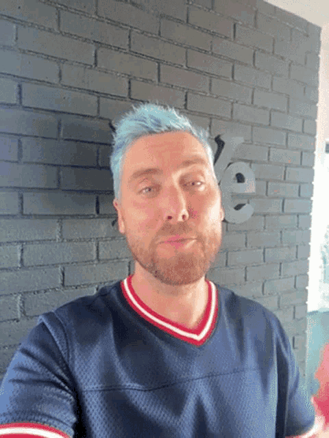 a man with blue hair and a beard is standing in front of a brick wall with the letter e on it