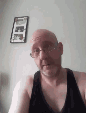 a bald man wearing glasses and a black tank top is sitting in front of a wall .