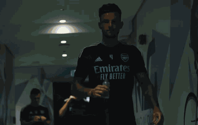 a man wearing a black emirates fly better jersey walking down a hallway