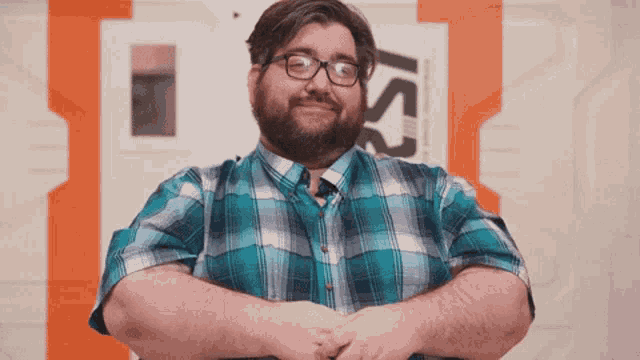 a man wearing glasses and a plaid shirt stands with his arms crossed in front of a sign that says is2