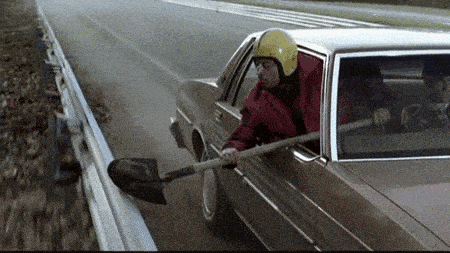 a man in a helmet is driving a car with a shovel sticking out the window .
