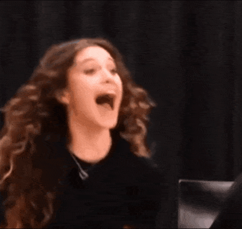 a woman with long curly hair is laughing with her mouth open and a hand behind her .