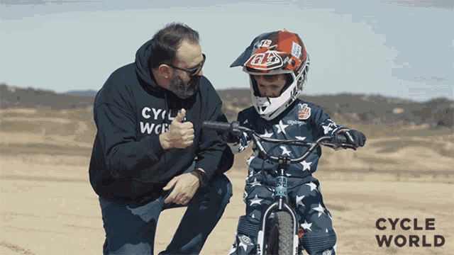 a man kneeling next to a child wearing a helmet that says cycle world on it