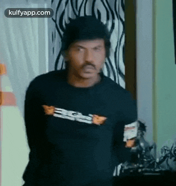 a man with a mustache is standing in a room wearing a black shirt .