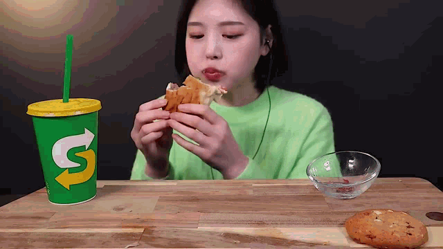 a woman in a green sweater is eating a sandwich next to a subway cup