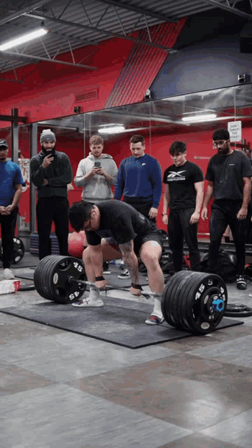 a group of men are watching a man lift a barbell with a reebok shirt on