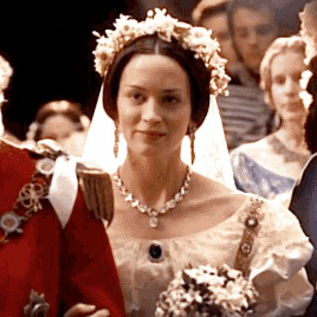 a woman wearing a veil and a necklace stands next to a man in a red jacket