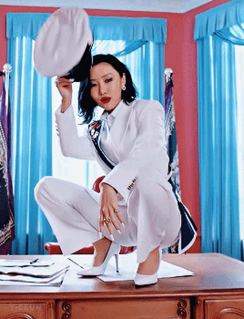 a woman in a white suit is squatting on a desk and holding a hat