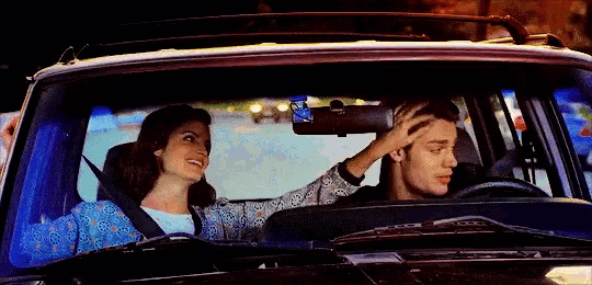 a man and a woman are driving a car and the woman is touching the man 's head