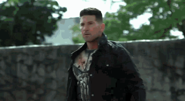 a man wearing a black jacket and a t-shirt with the punisher on it stands in front of a stone wall