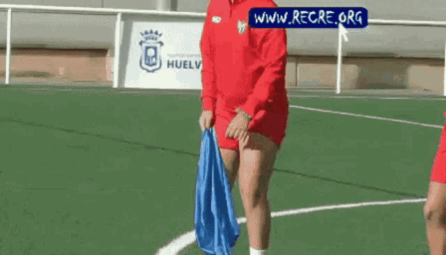 a woman in a red shirt is standing on a soccer field holding a blue bag .