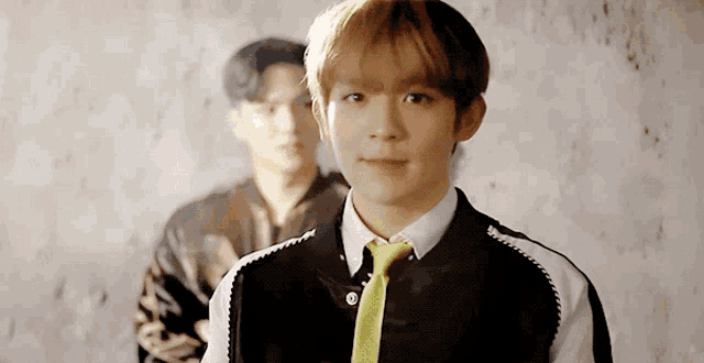 a young man wearing a black jacket and a yellow tie stands in front of a wall .