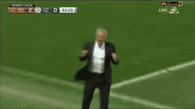 a man in a suit stands on a soccer field in front of a scoreboard that says premier league live