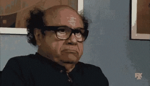 a bald man wearing glasses and a black shirt is sitting in front of a wall .