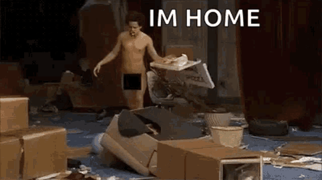 a shirtless man in underwear is standing in a room filled with boxes and a sign that says im home .