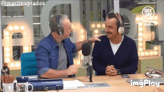 a man wearing headphones talks to another man in front of a microphone with a dee jay mug on the table