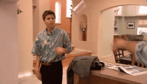a man in a colorful shirt is standing in a living room next to a table with a magazine on it .