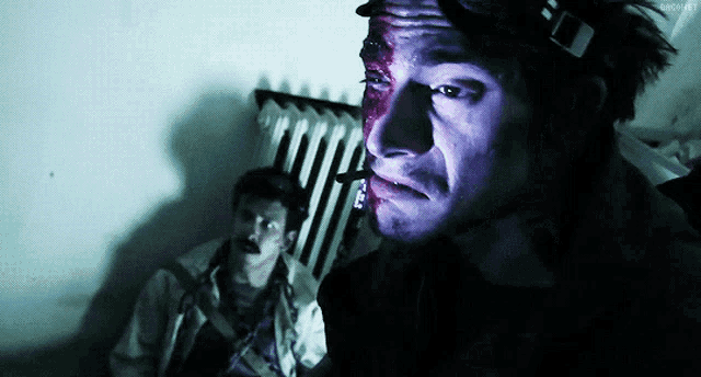 a man with blood on his face is smoking a cigarette while another man looks on