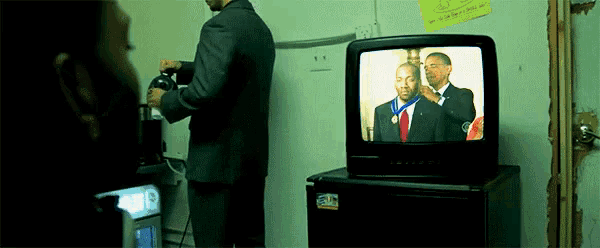 a man in a suit stands in front of a television showing barack obama receiving a medal