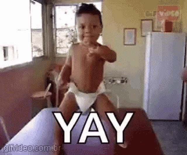 a baby in a diaper is standing on a table and pointing at the camera with the word yay above him