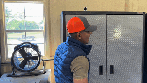 a man wearing an orange hat stands in front of a fan in a garage