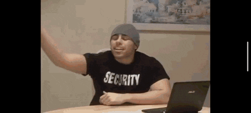 a man wearing a security shirt and a beanie is sitting at a table with a laptop .