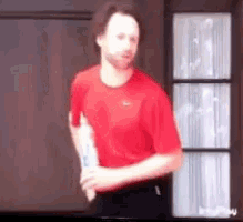 a man in a red shirt is standing in front of a window holding a bottle .